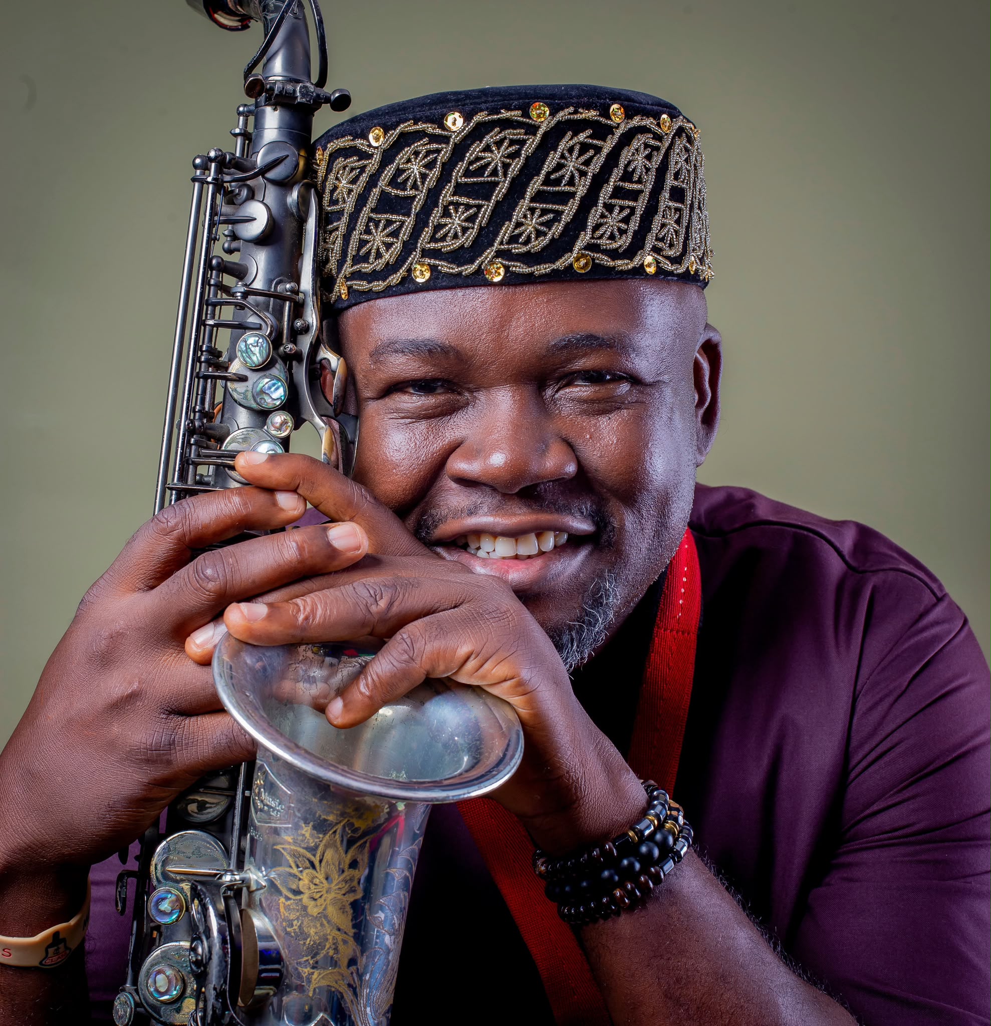 Segun Oluwayomi holding saxophone in royal embroidered Yoruba cap and burgundy agbada